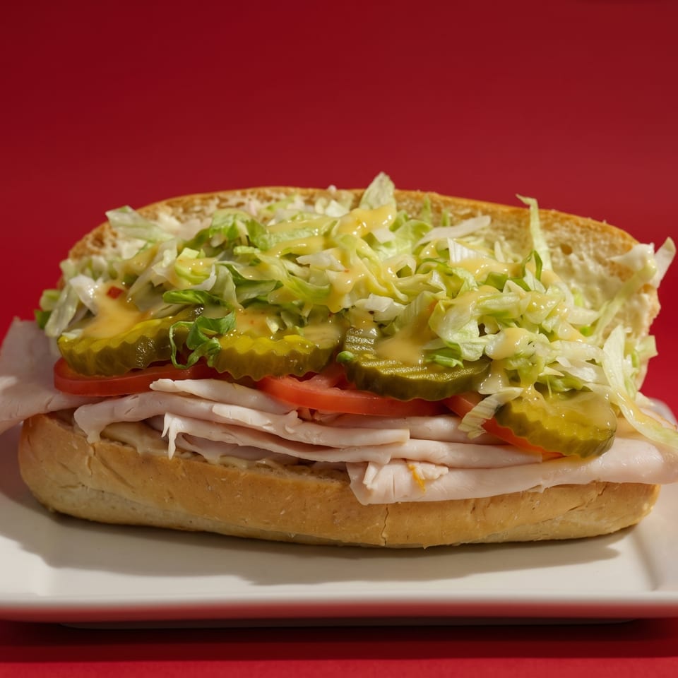 Turkey sub sandwich with lettuce, pickles, tomatoes, and sauce on a white plate against a red background.
